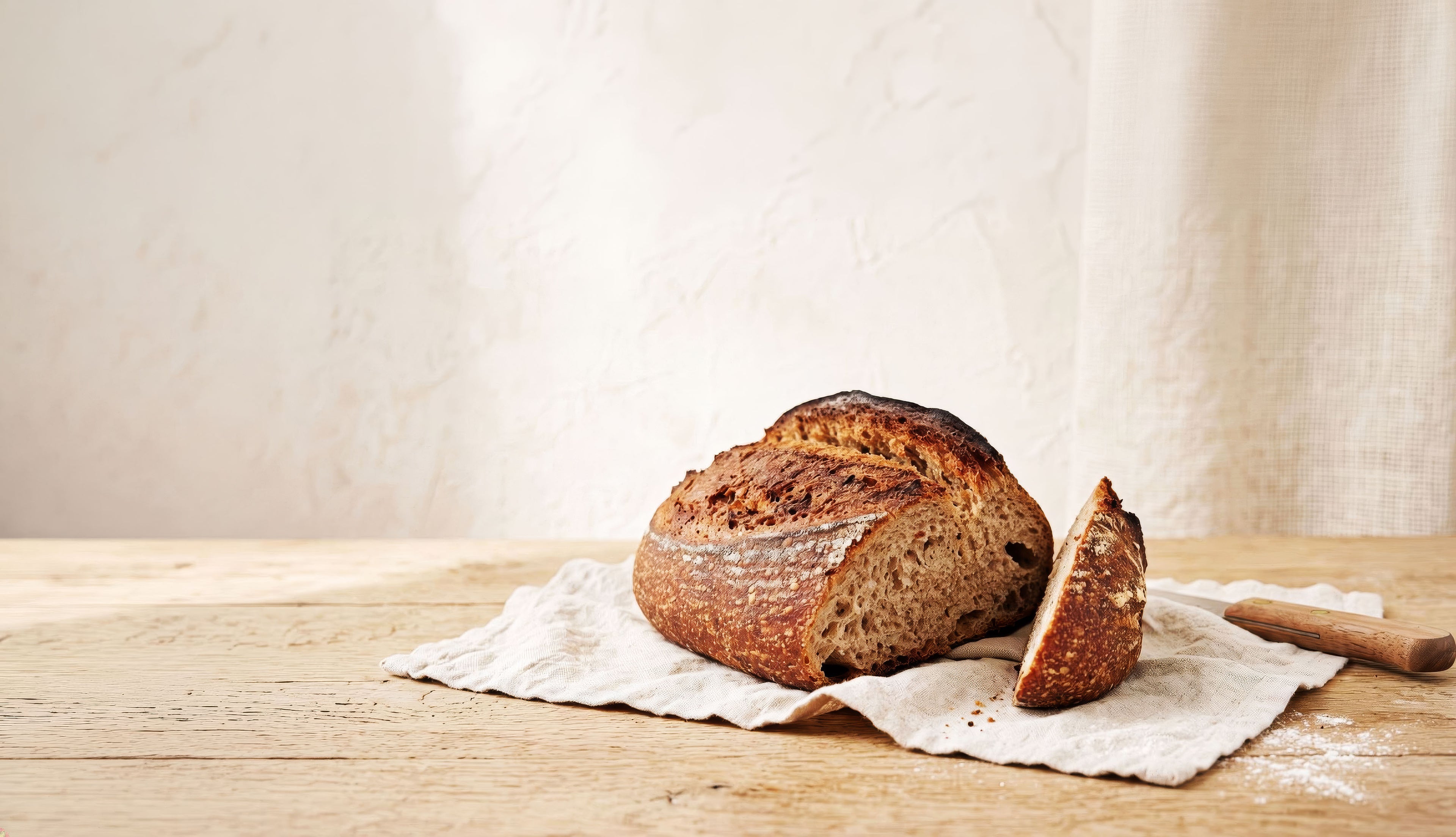 Zubi Bakes wholewheat sourdough loaf with a golden-brown crust sits on cream bakers linen on a wooden table, with a slice cut and placed beside it.